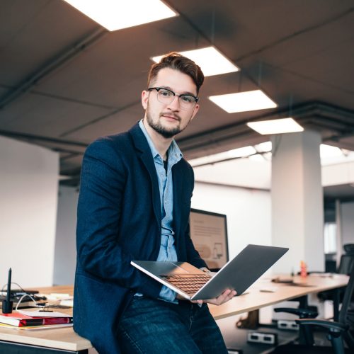 Attractive man in glassess is sitting near the workplace in office. He wears  blue shirt, dark jacket.  He holds laptop and looks to the camera.